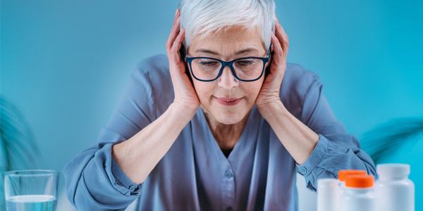 Person sitzt an einem Tisch und hält mit beiden Händen den Kopf. Auf dem Tisch stehen mehrere Behälter sowie ein Glas Wasser.